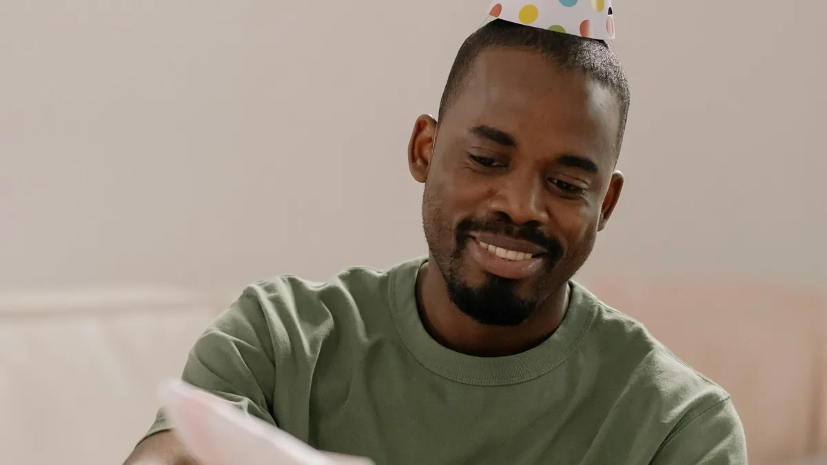 Smiling man in a party hat opening a birthday reveal card in a birthday setting.