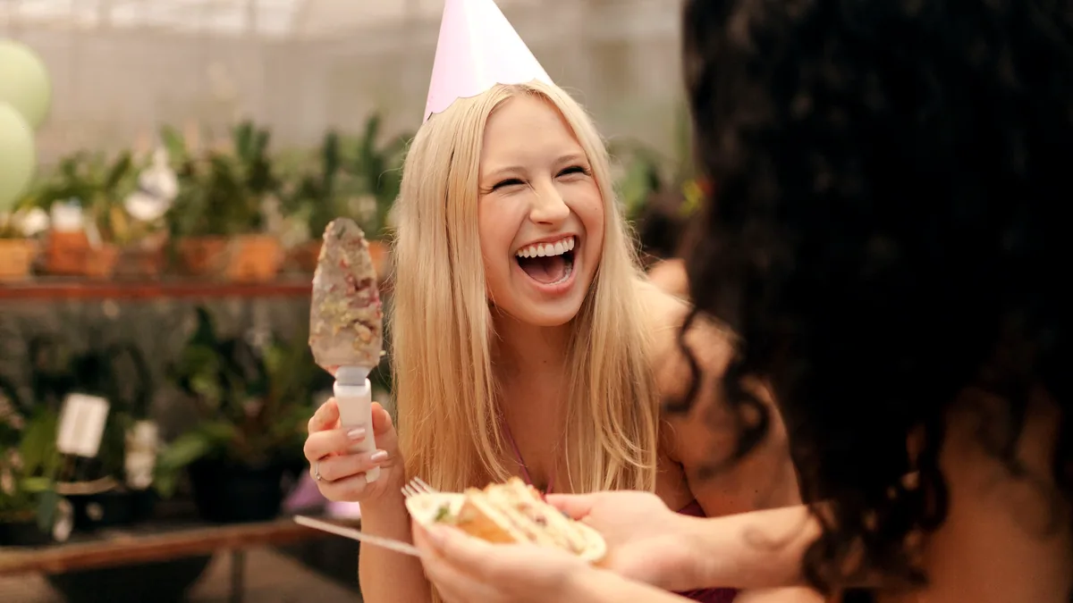 Two friends laughing during a birthday surprise with cake and party hats.