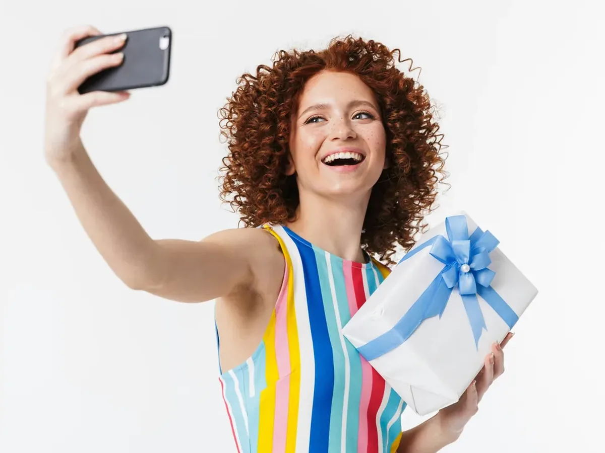 A smiling woman holding a phone and wrapped gift for an evergreen surprise gift reveal.